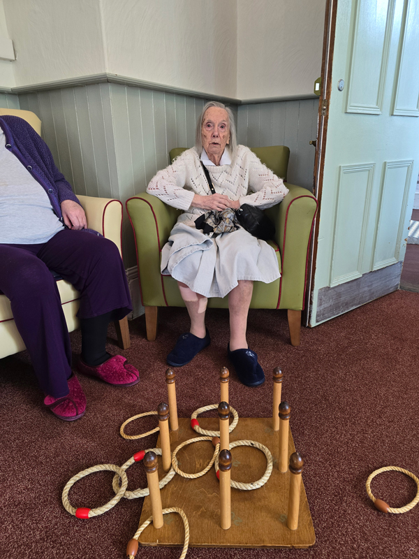 Barbara seated having a go throwing rope rings onto a board of wooden pegs on the floor