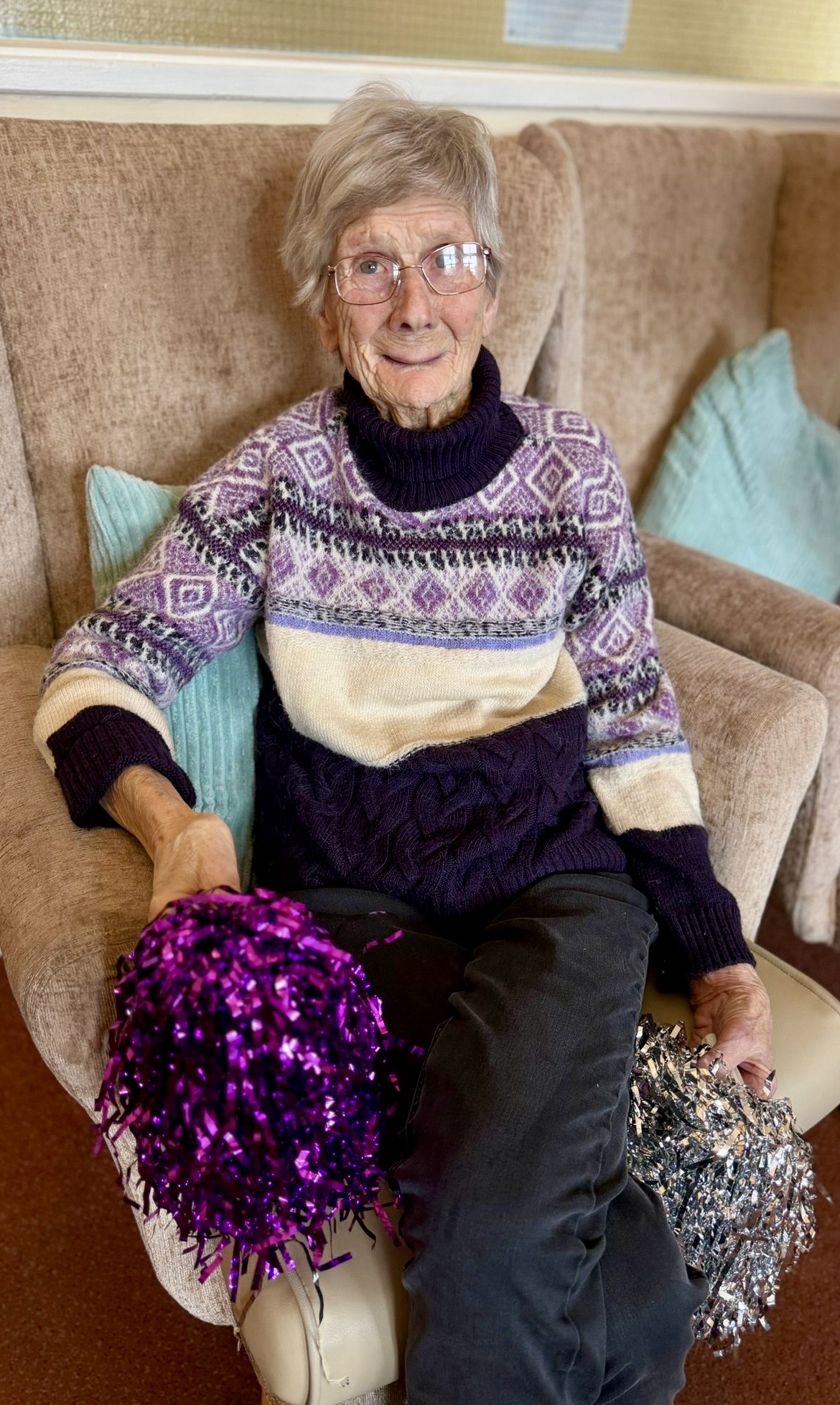 Female resident seated holding a purple and a silver shiny pompoms smiling