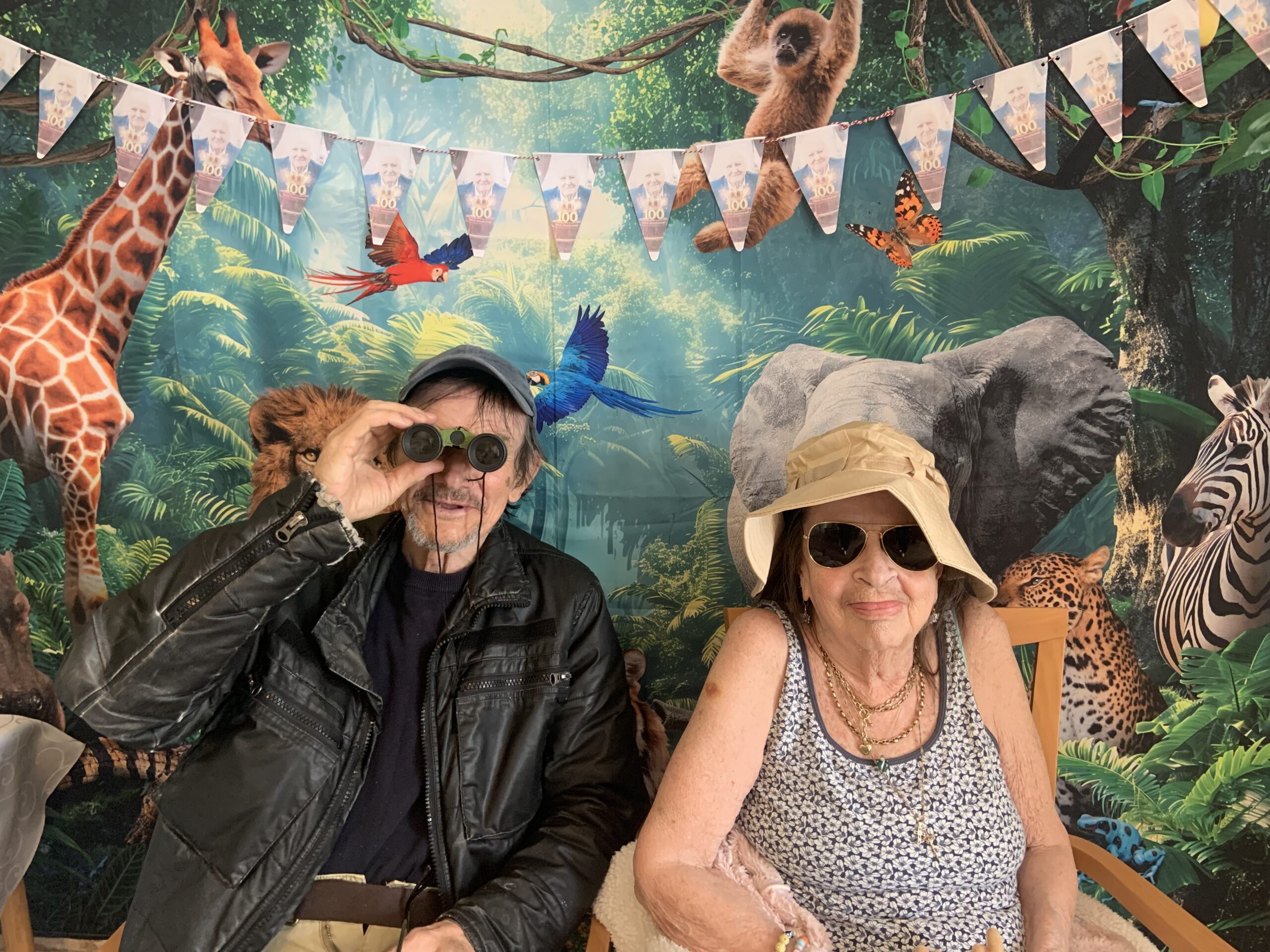 Male resident holding binoculars seated with a female resident in front of a jungle back drop scaled
