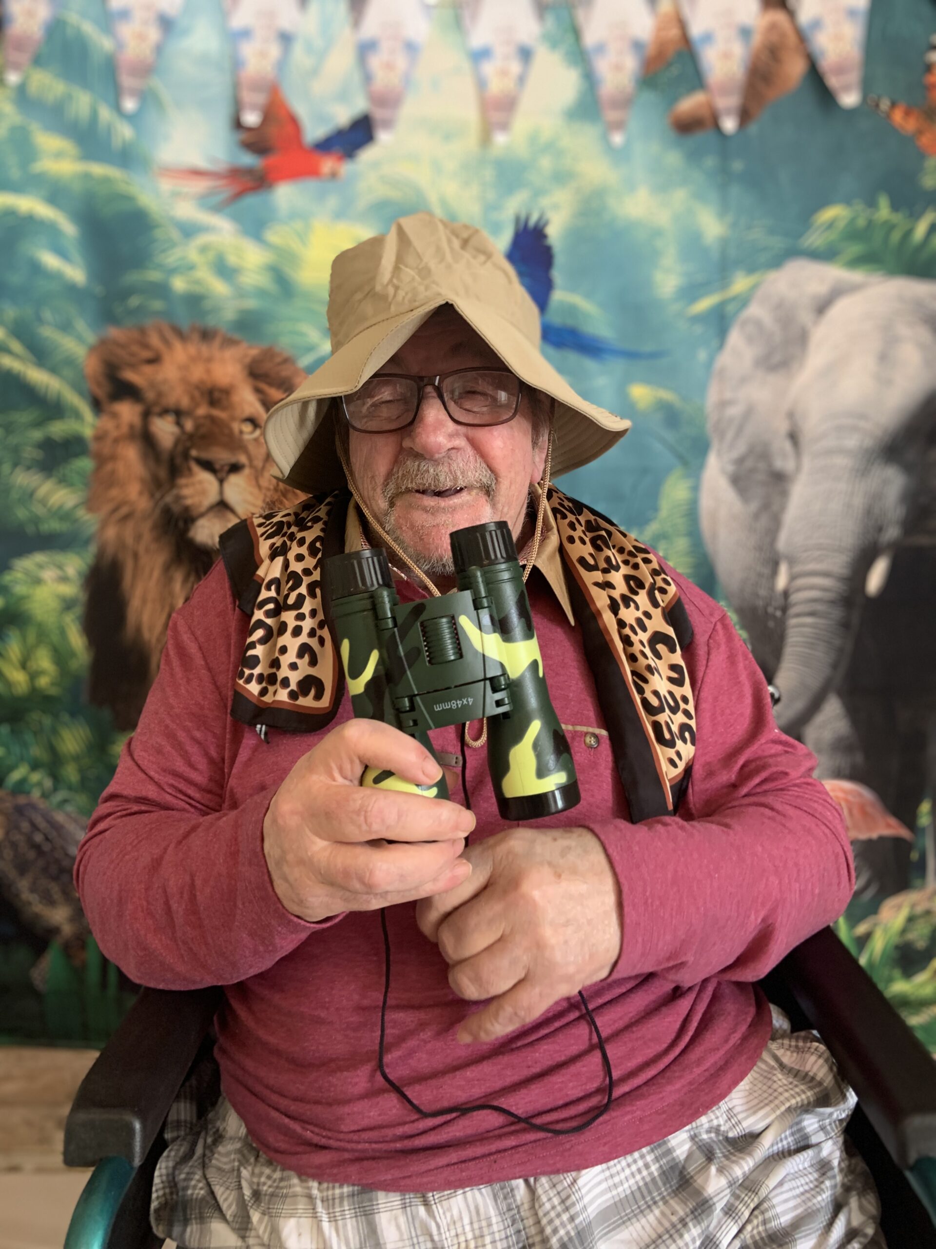Male resident in a safari hat with binoculars seated in front of jungle back drop scaled