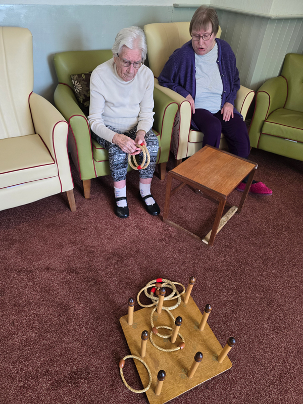 Shirley seated having a go throwing rope rings onto a board of wooden pegs on the floor while Chris watches